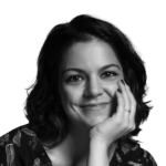Retrato a preto e branco de Joana Castro, a sorrir, apoiada com o queixo na mão.